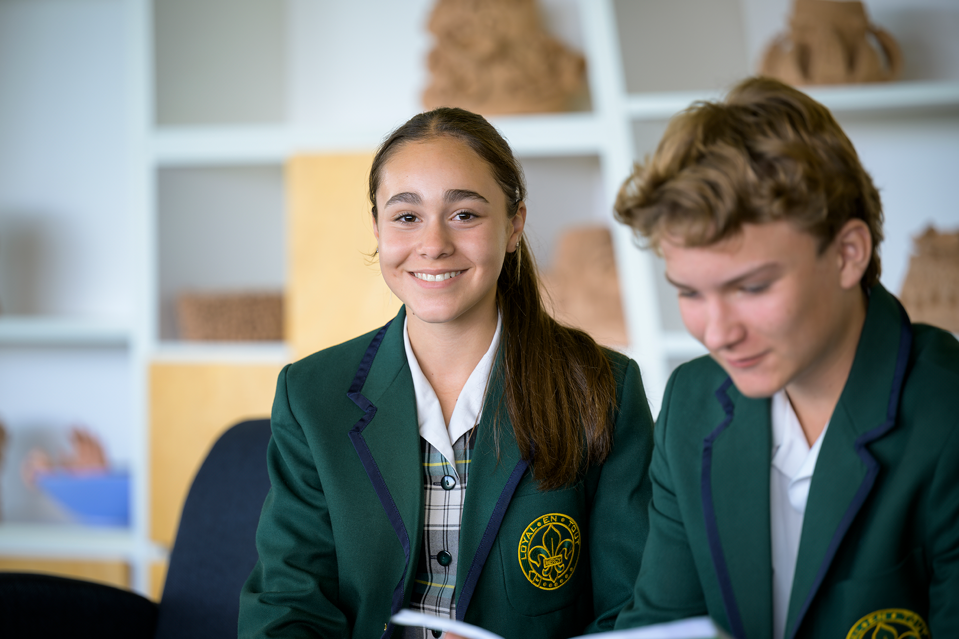 Smiling student in a green school blazer sitting beside a classmate in a classroom setting.